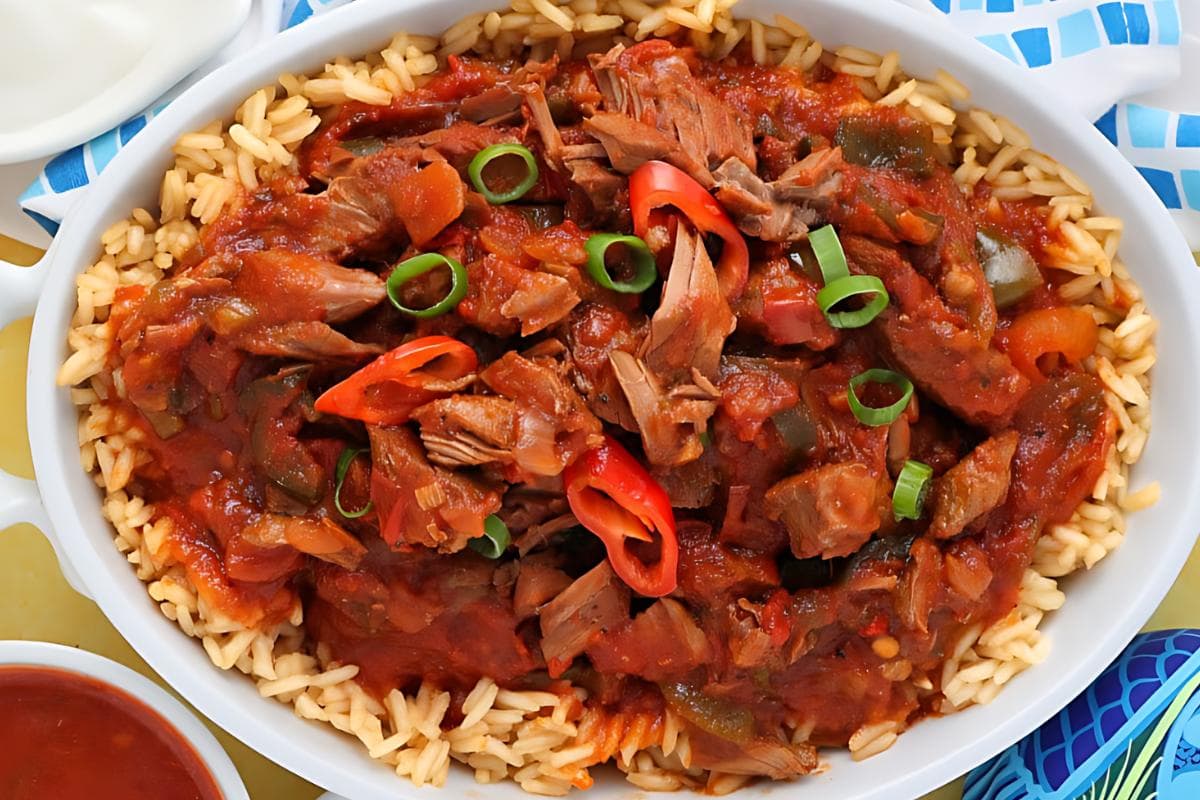 A white dish filled with rice topped with shredded meat in a tomato-based sauce, garnished with sliced red chili and green onions.