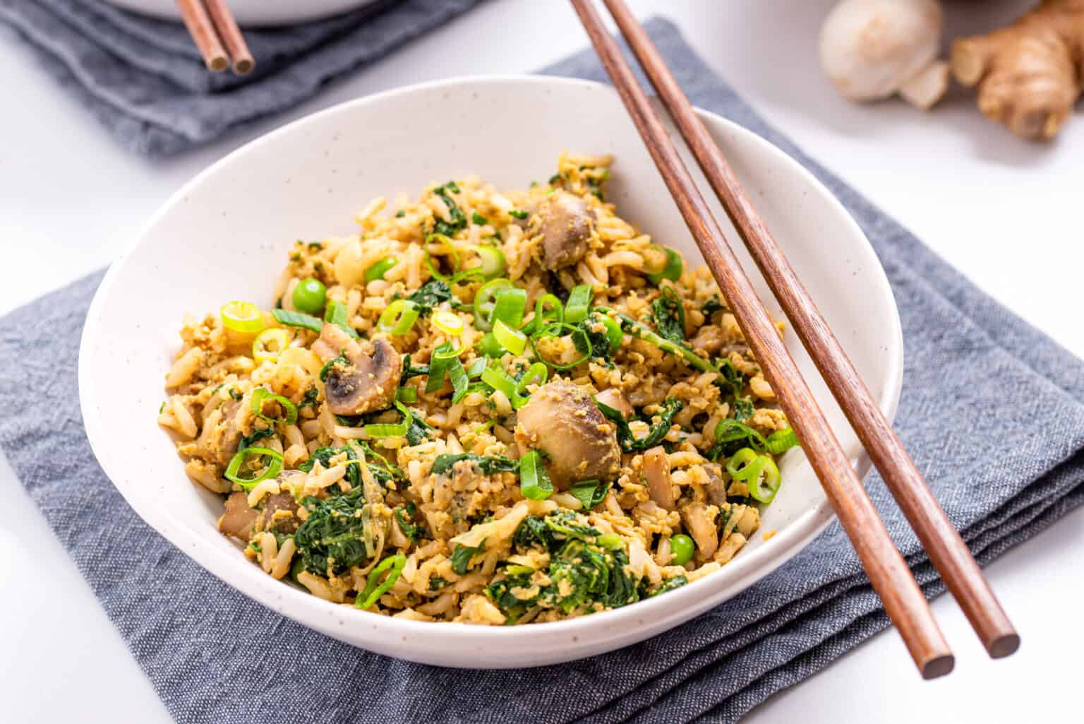 A white bowl filled with fried rice, mushrooms, spinach, and chopped green onions, with wooden chopsticks resting on the bowl.