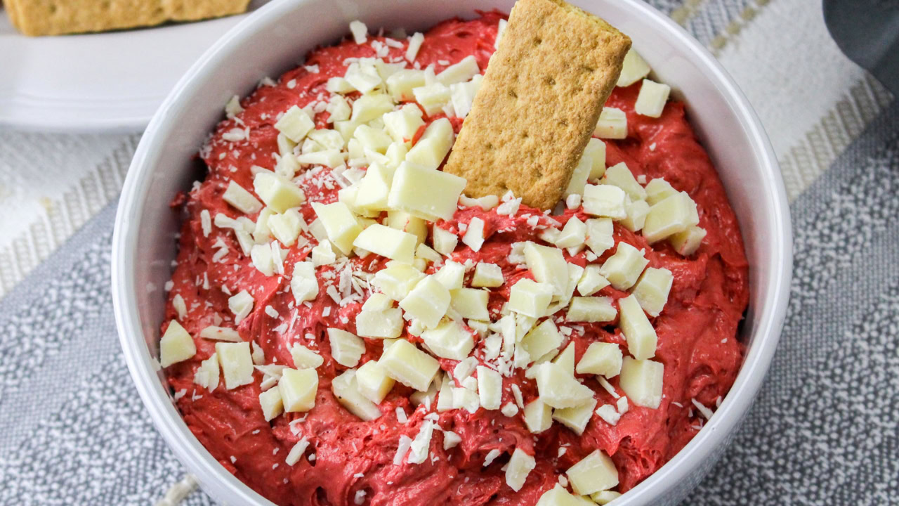 A bowl of red dip topped with white chocolate chunks and a rectangular graham cracker sticking out.