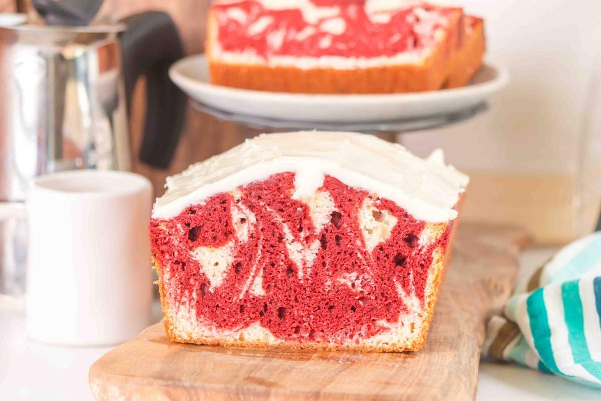 A sliced loaf of red and white marbled cake with white frosting sits on a wooden board; more cake is visible in the background.