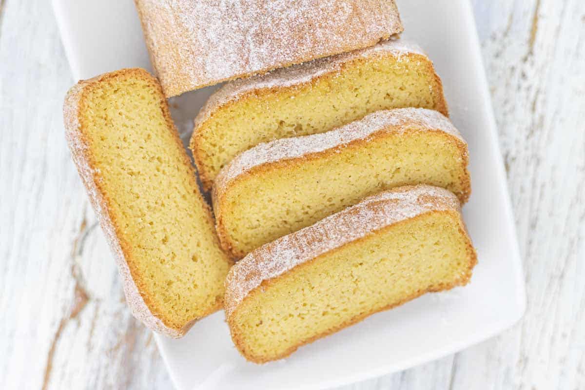 Four slices of a golden, powdered sugar-dusted pound cake are arranged on a white rectangular plate.