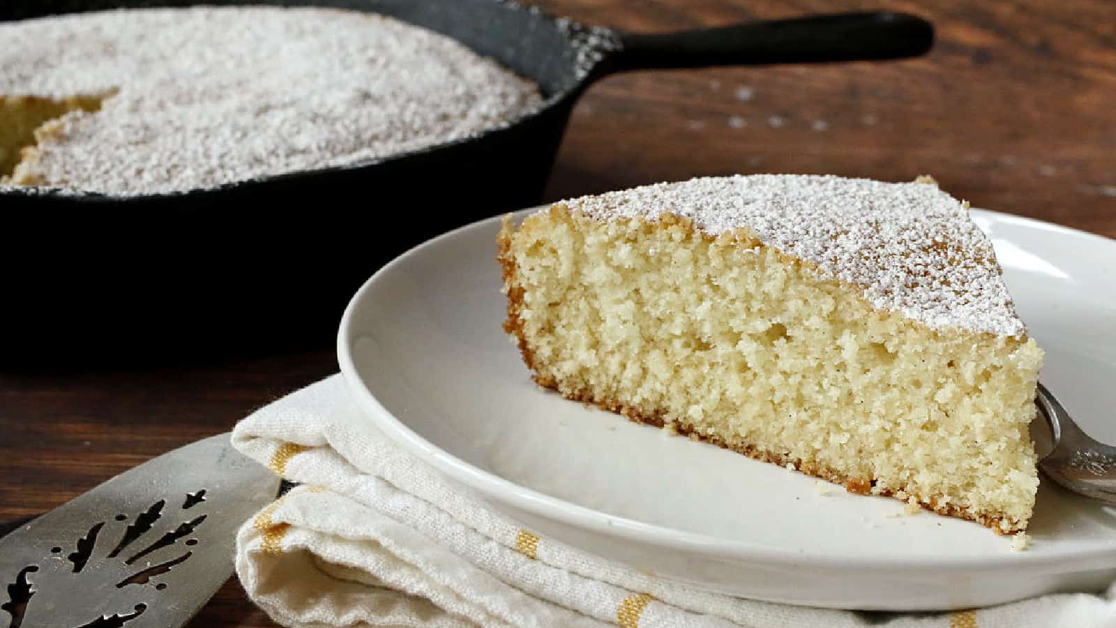 A slice of powdered sugar-topped cake on a white plate with a fork, next to a napkin; the rest of the cake is in a cast iron skillet in the background.