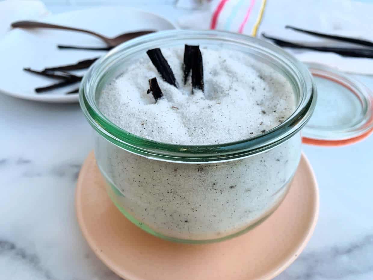 A glass jar filled with vanilla sugar, topped with vanilla bean pieces, sits on a peach plate with a lid and more vanilla beans in the background.