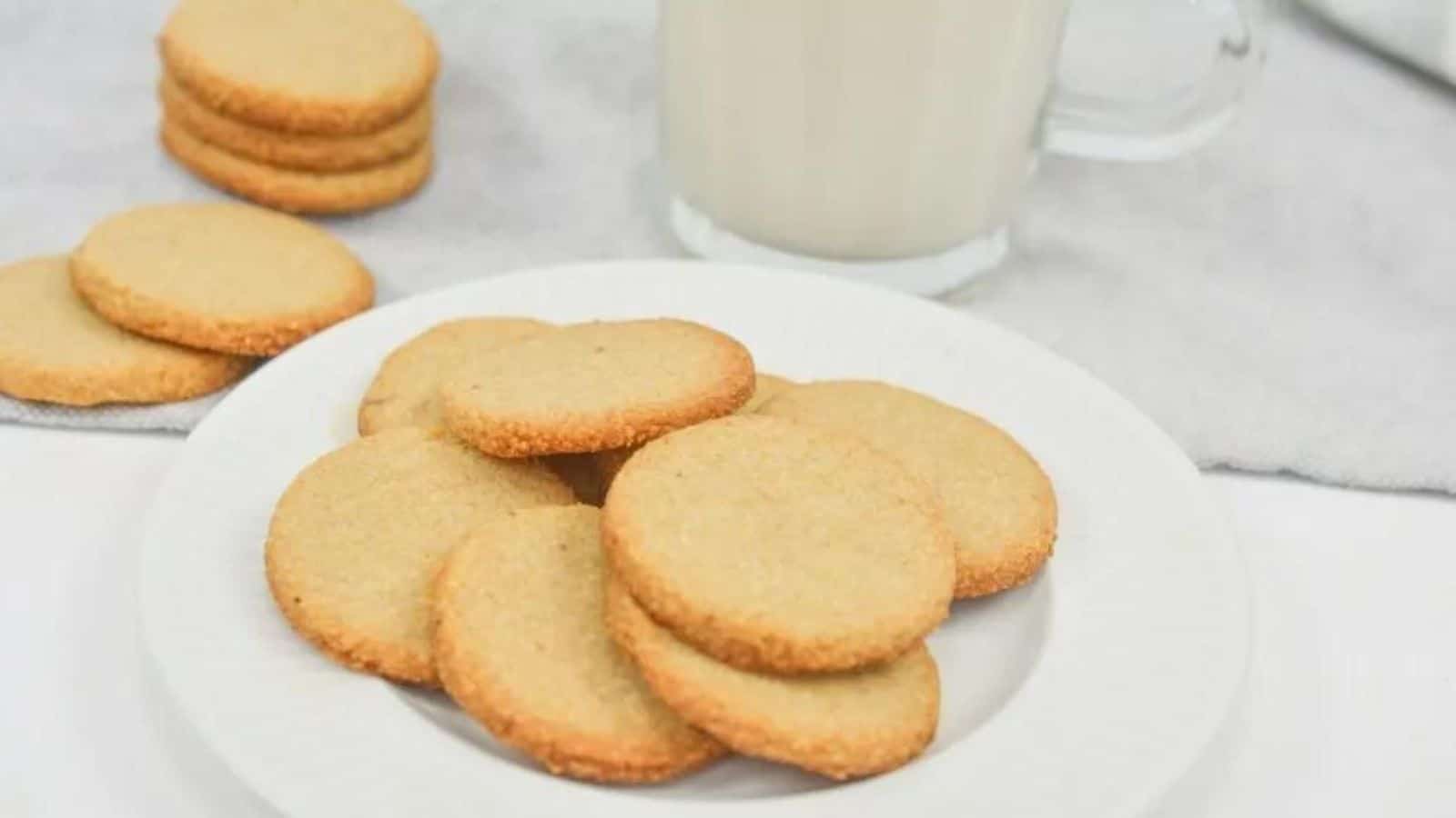 A white plate with several round, plain cookies is placed in front of a glass of milk, with more cookies stacked in the background.