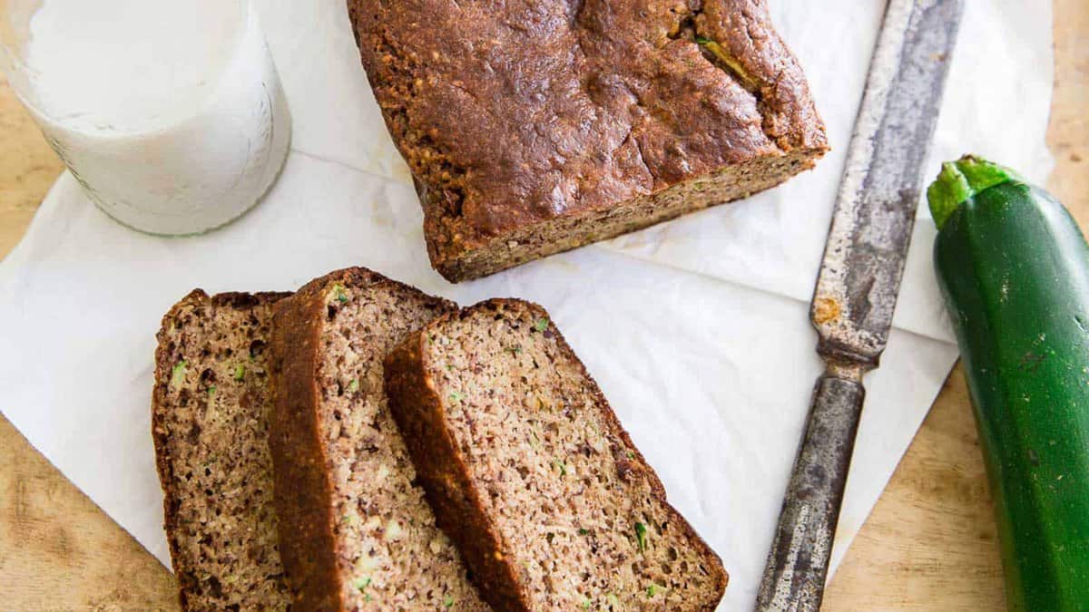 A loaf of zucchini bread with two slices cut, a glass of milk, a zucchini, and a knife on a parchment paper-lined wooden surface.