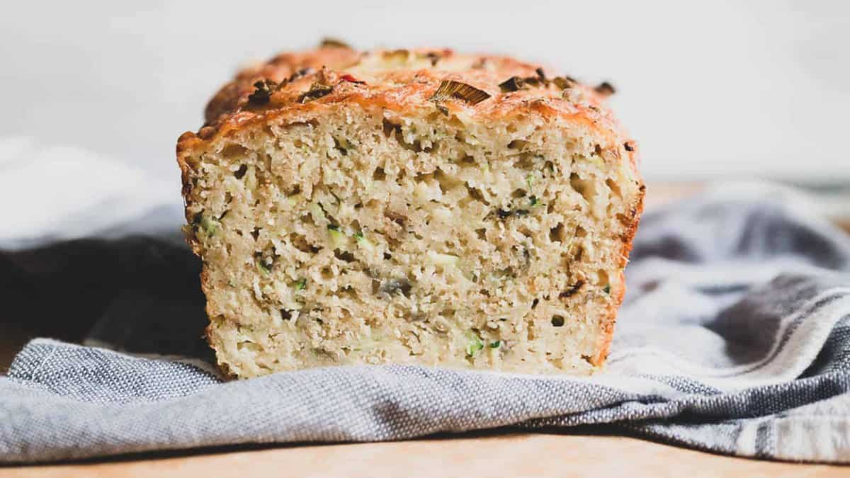 A close-up of a sliced loaf of savory bread with visible herbs and vegetables, resting on a gray cloth.