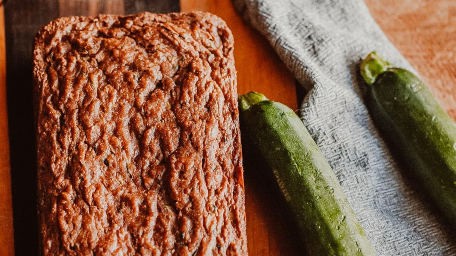 A loaf of zucchini bread sits on a wooden surface next to two whole zucchinis and a folded gray dish towel.