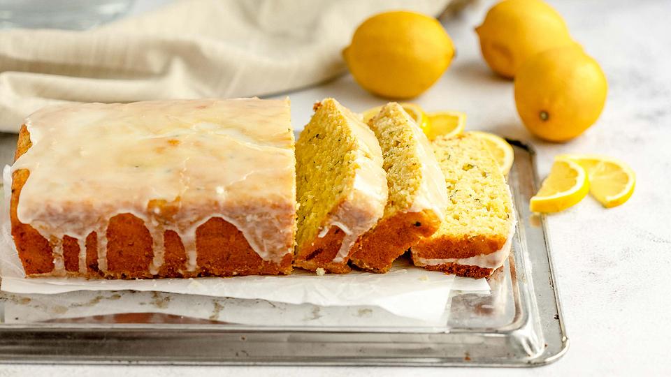 A glazed lemon loaf cake sits on a tray, partially sliced, with whole lemons and lemon wedges in the background.