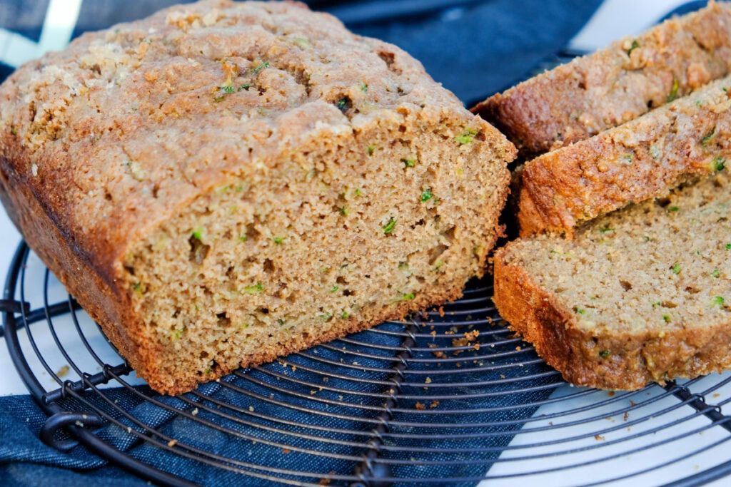 A loaf of zucchini bread with a golden-brown crust, partially sliced, sits on a round cooling rack.