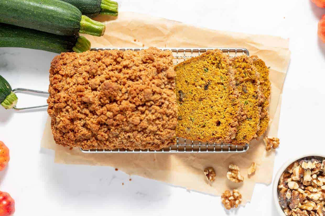 A loaf of zucchini bread with crumb topping on a cooling rack, partially sliced, surrounded by fresh zucchini and walnuts on a parchment-lined surface.