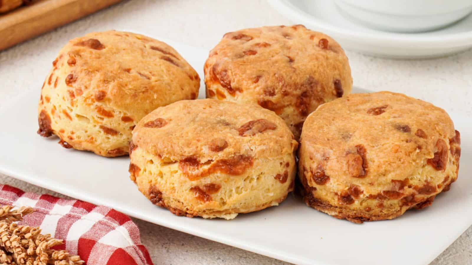 Four golden brown cheese scones are arranged on a white rectangular plate, with a red and white checkered cloth and a cup in the background.