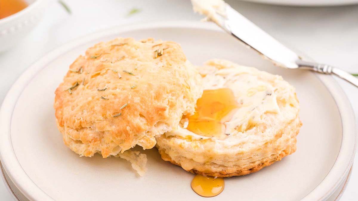 A biscuit split open on a plate, spread with butter and drizzled with honey, with a butter knife resting nearby.