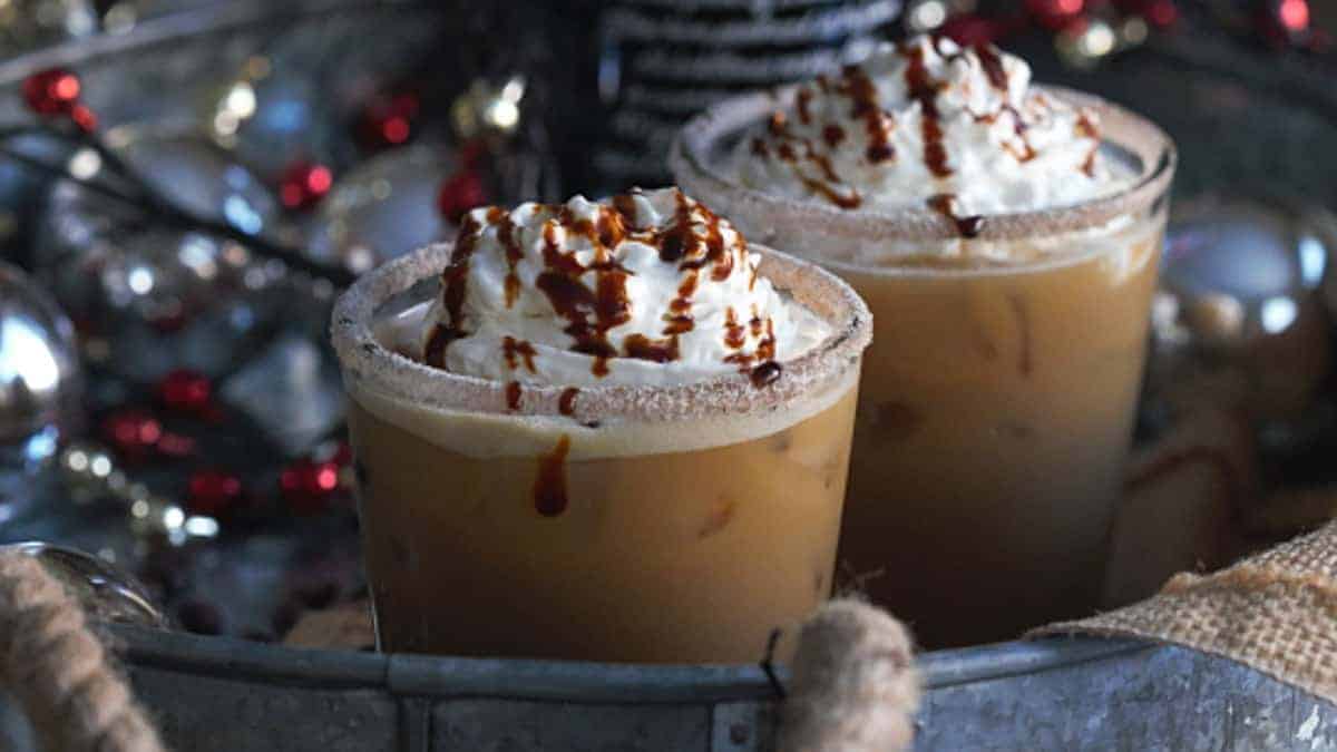 Two iced coffee drinks topped with whipped cream and chocolate drizzle, served in glasses with sugared rims, set among festive holiday decorations.