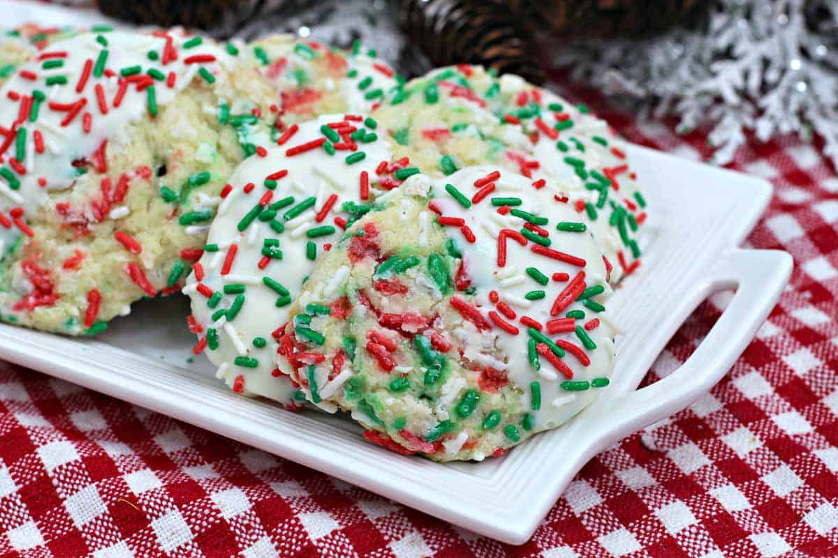 Three cookies topped with white icing and red and green sprinkles on a white rectangular plate, placed on a red checkered cloth.