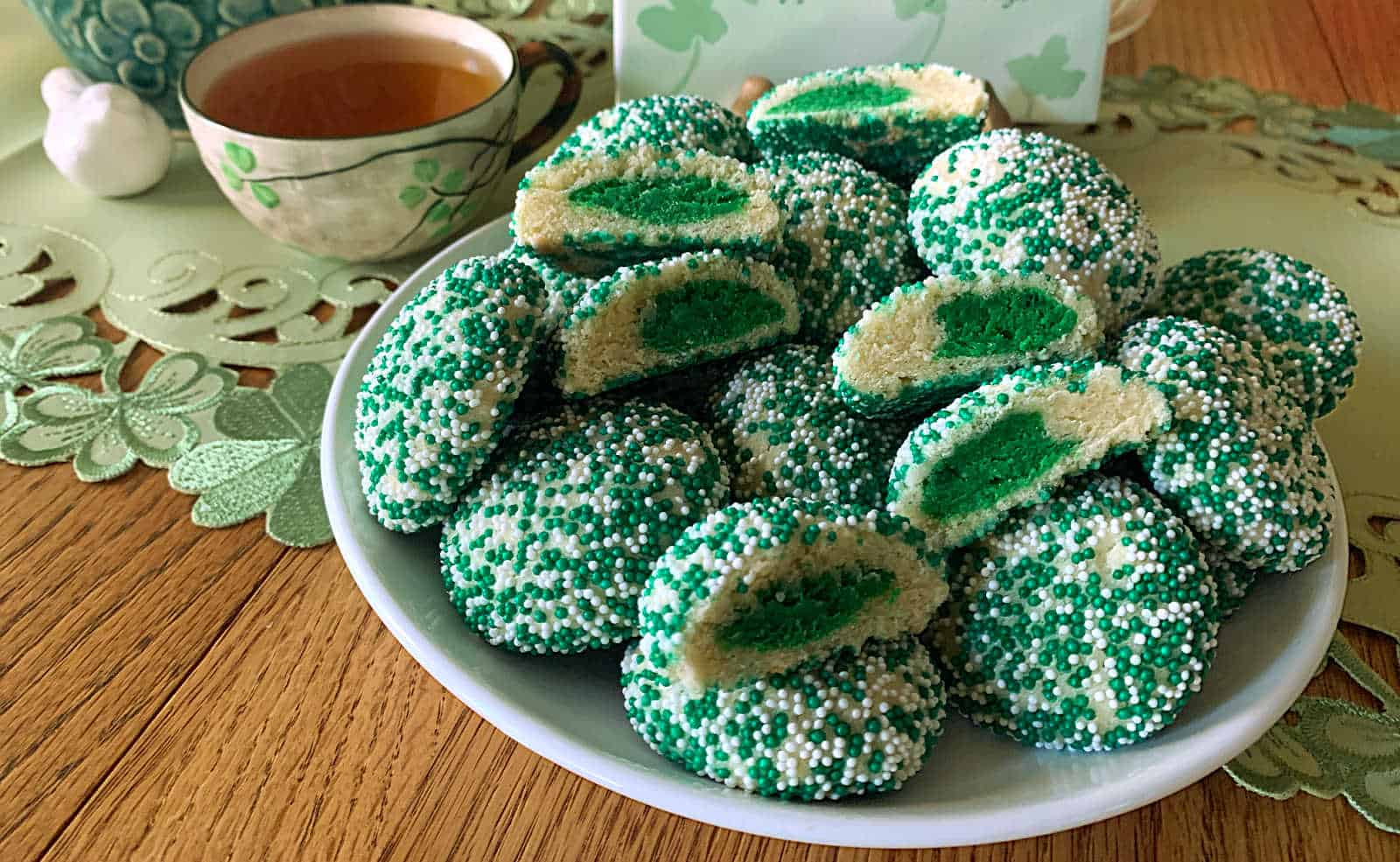 A plate of round green and white sprinkle-coated cookies with green centers sits on a wooden table next to a teacup.