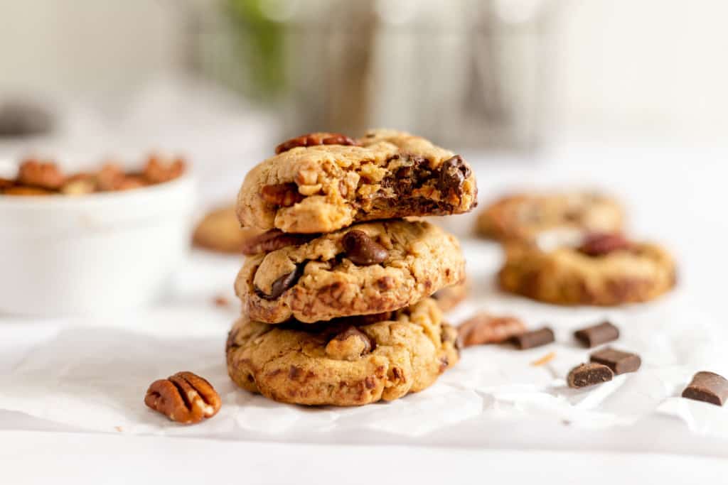 Three chocolate chip pecan cookies stacked on parchment paper, with the top cookie partially eaten. Chocolate chunks and pecans are scattered nearby. A bowl of pecans is in the background.