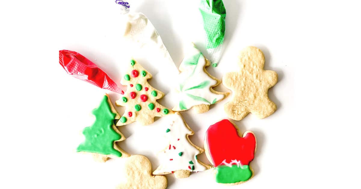 Assorted undecorated and decorated Christmas cookies in tree, mitten, and gingerbread shapes with piping bags of red, white, and green icing on a white background.