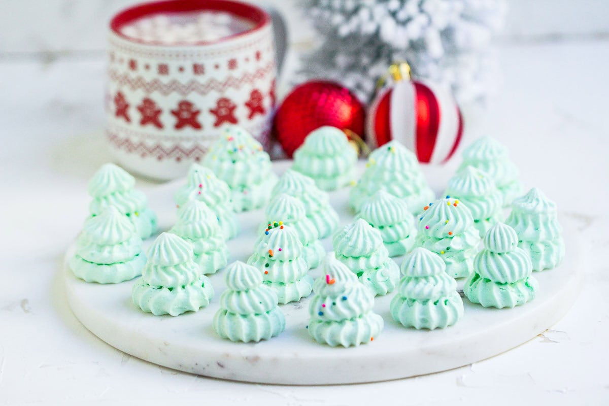 Light green meringue cookies shaped like Christmas trees with sprinkles on a white plate; red ornaments and a festive mug in the background.
