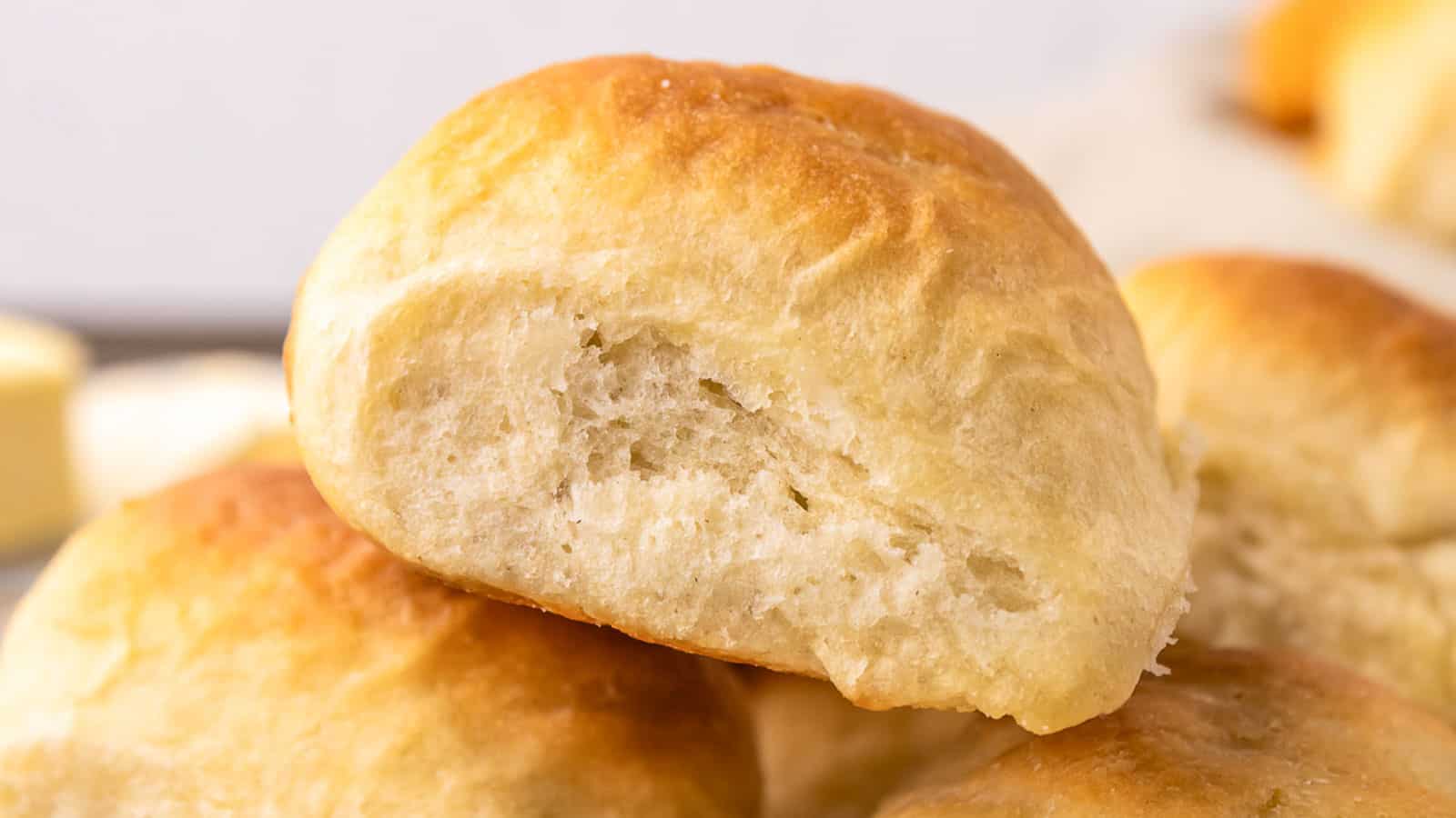 A close-up of a stack of golden brown dinner rolls, with one roll showing a bite taken out, revealing its soft, fluffy interior.