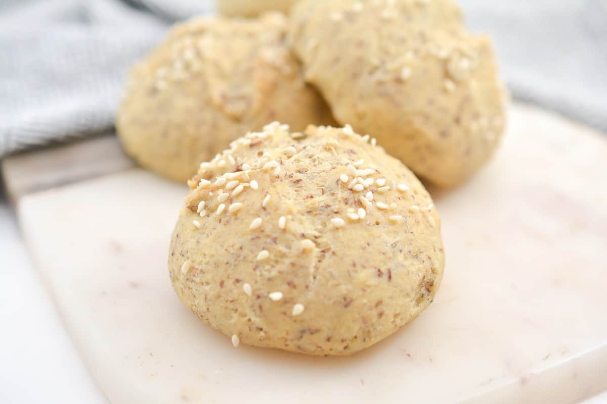 A close-up of a round bread roll topped with sesame seeds, resting on a white marble surface with more rolls in the background.