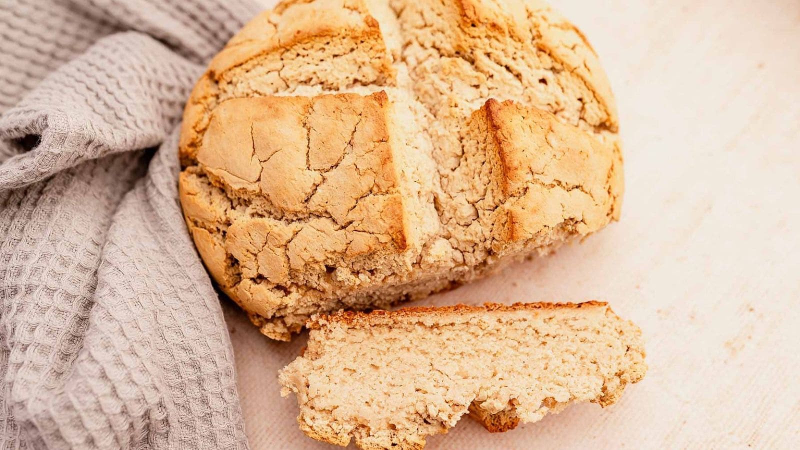 A round loaf of rustic bread with a cracked crust sits on a light surface next to a gray textured cloth; one thick slice is cut and placed in front.