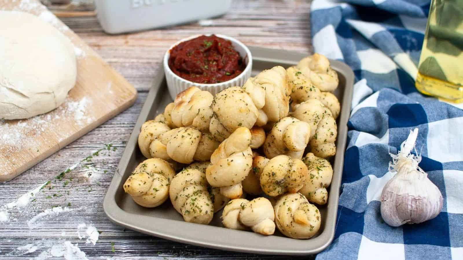 A baking tray filled with garlic knots garnished with herbs, a ramekin of marinara sauce, pizza dough, olive oil, and garlic on a wooden table.
