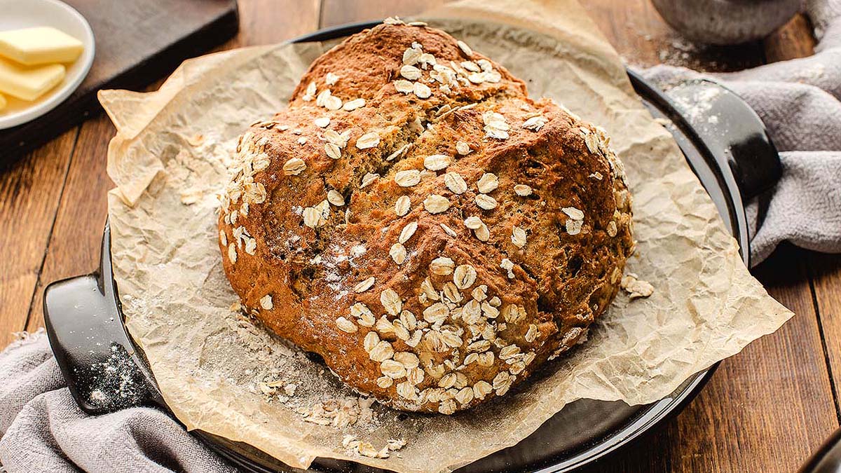 A round loaf of oat-topped brown bread sits on parchment paper in a black baking dish on a wooden table.