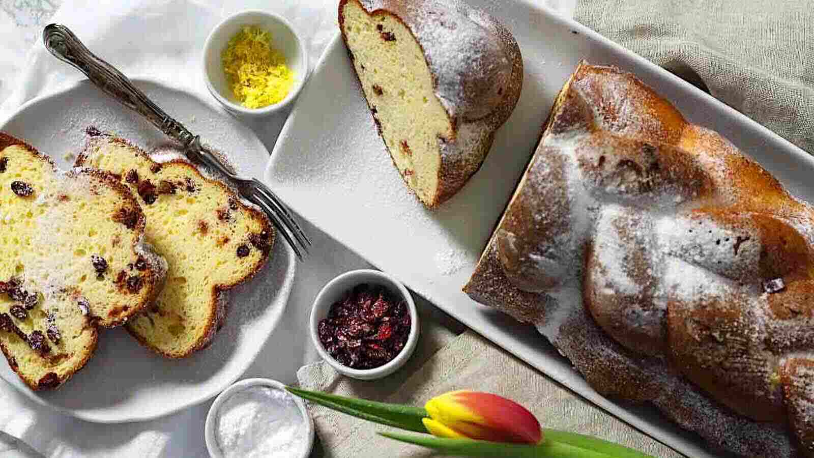 A sliced loaf of sweet bread with dried fruit, dusted with powdered sugar, is served on a white platter with lemon zest, dried cranberries, and a yellow-red tulip on the side.