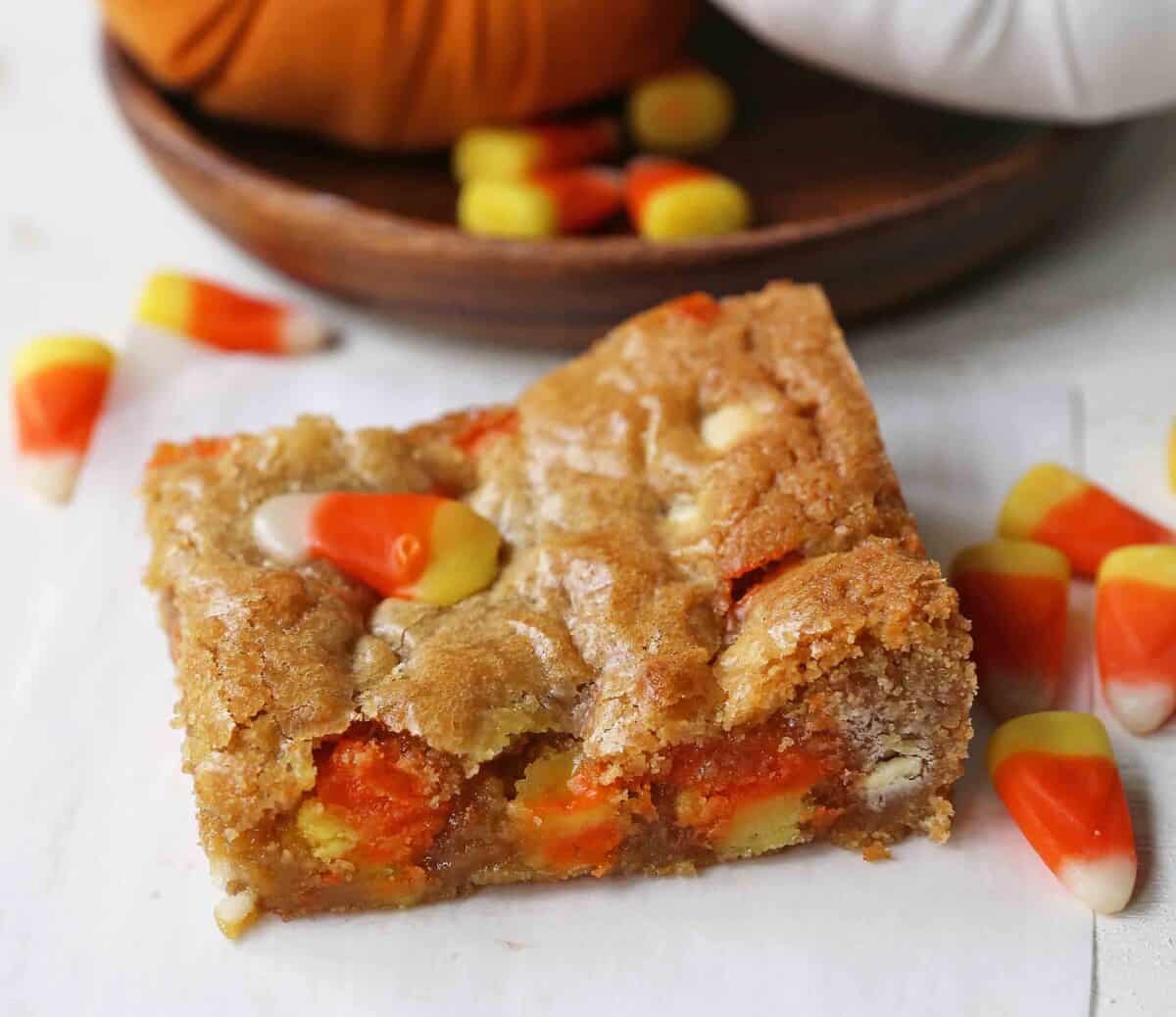 A blondie bar with candy corn pieces is placed on a white surface, with more candy corn scattered nearby and a wooden plate in the background.