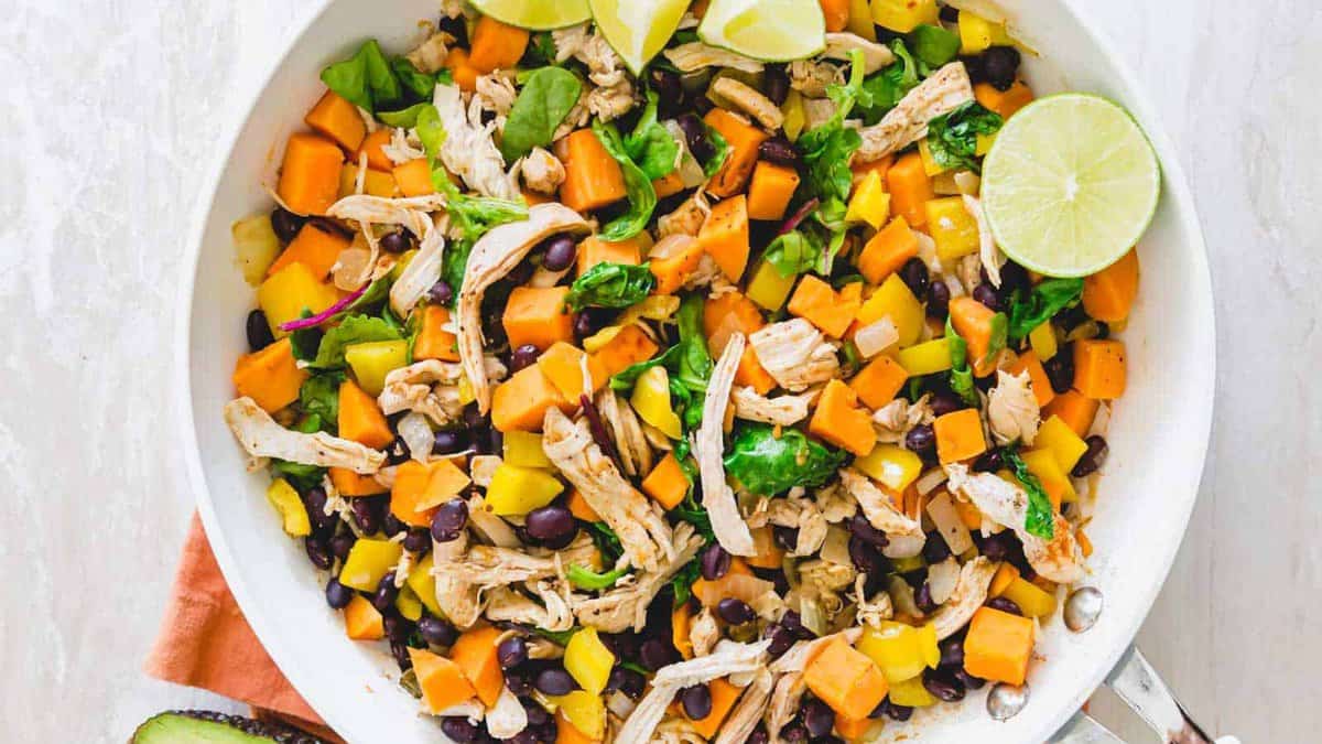 A white skillet filled with shredded chicken, black beans, diced sweet potatoes, spinach, and lime wedges.