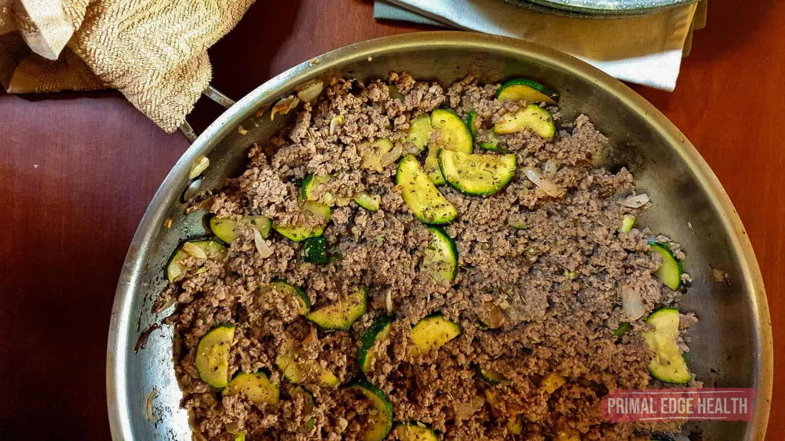 A skillet filled with cooked ground beef, sliced zucchini, and onions on a wooden surface.