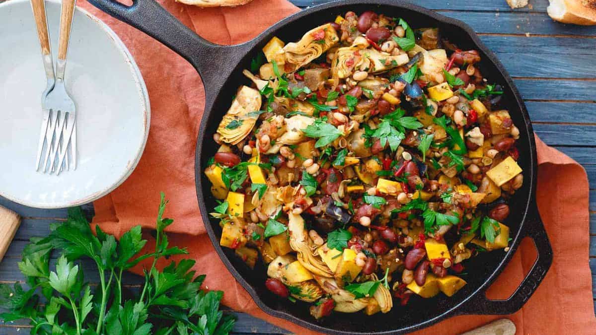 A cast iron skillet filled with a cooked vegetable and bean medley, garnished with chopped parsley, sits on a wooden table beside a plate, fork, and fresh parsley.