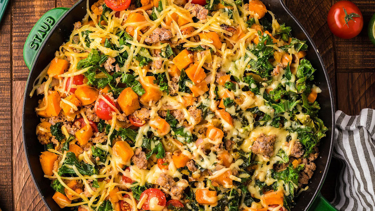 A skillet filled with a baked dish containing chopped kale, diced sweet potatoes, cherry tomatoes, ground meat, and melted cheese on top.