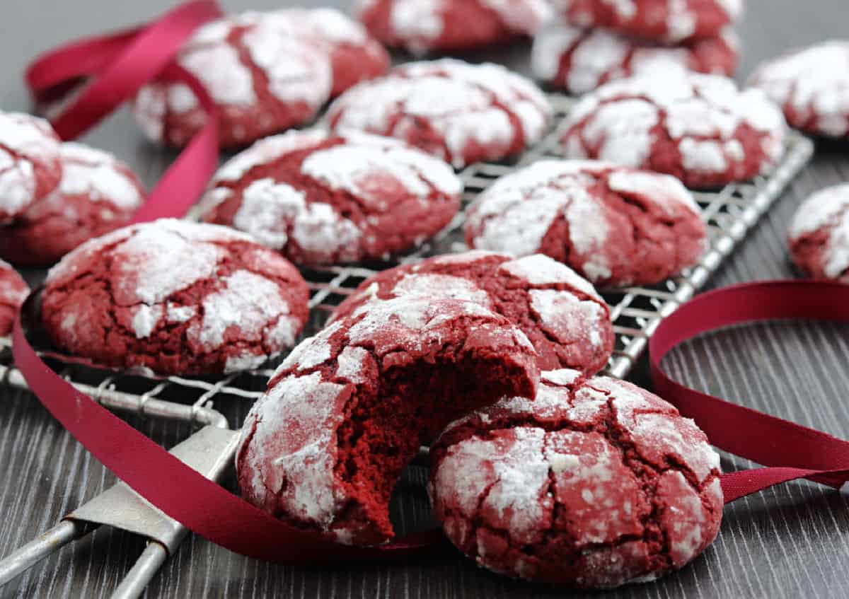 Red velvet crinkle cookies with powdered sugar on top are cooling on a rack; one cookie has a bite taken out of it, and a red ribbon is draped among the cookies.