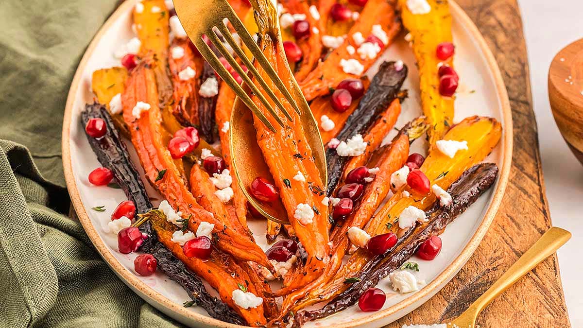 A plate of roasted multicolored carrots topped with crumbled cheese and pomegranate seeds, with a fork and knife slicing into a carrot.