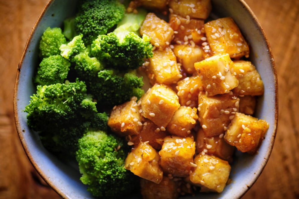 A bowl filled with steamed broccoli on one side and cubed tofu coated in sauce and sprinkled with sesame seeds on the other.