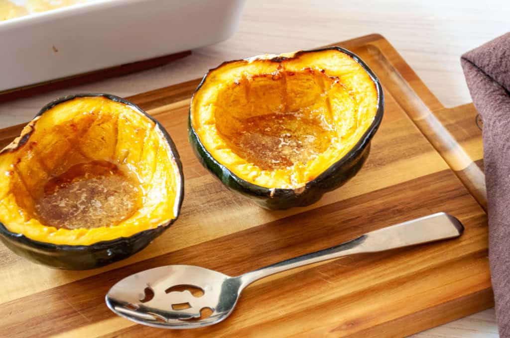 Two roasted acorn squash halves sit on a wooden cutting board next to a metal serving spoon and a brown napkin.