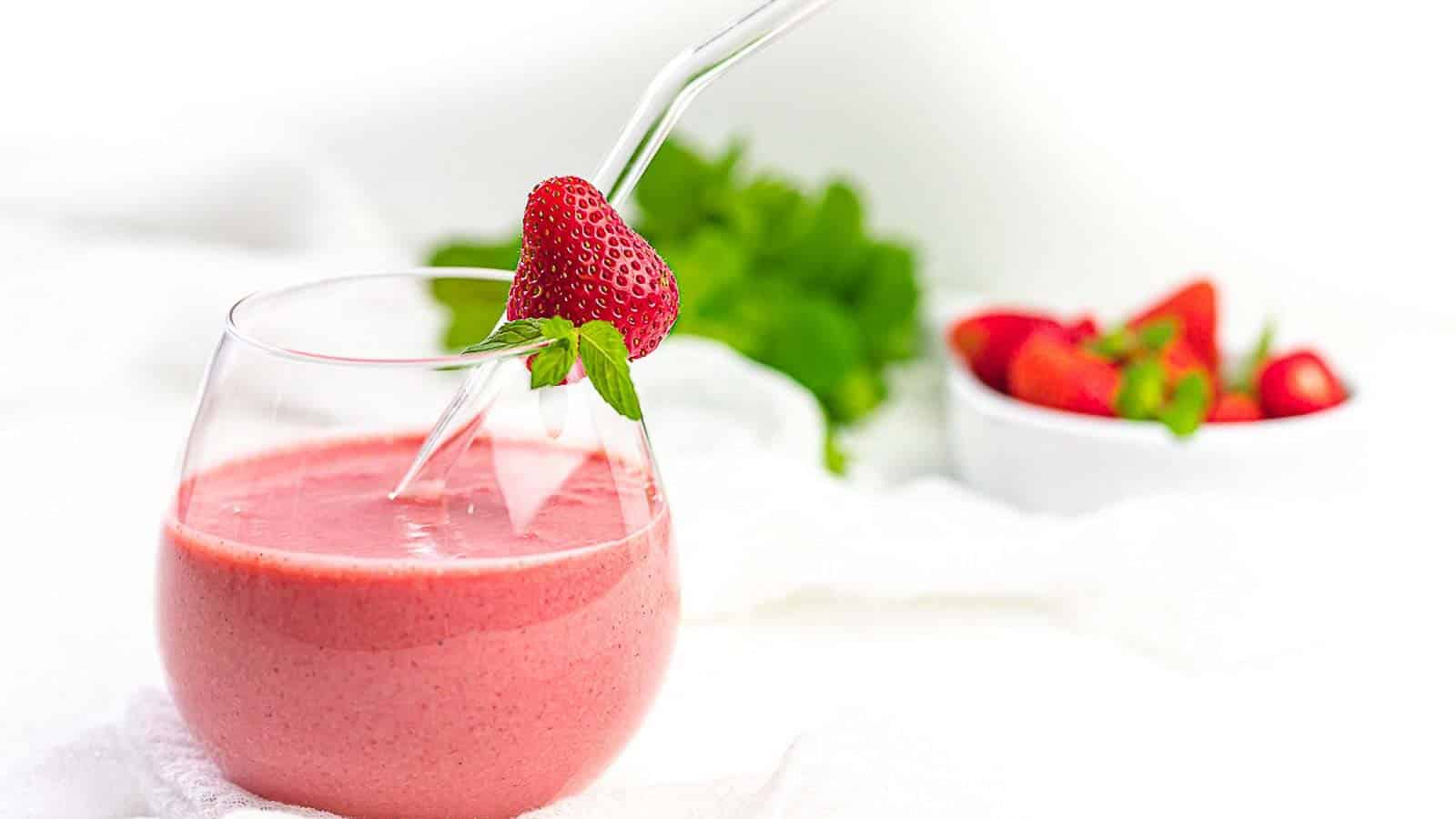 A glass of pink strawberry smoothie with a glass straw, garnished with a fresh strawberry; a bowl of strawberries is in the background.