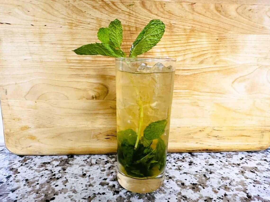 A tall glass filled with a light-colored drink, crushed ice, mint leaves, and a mint sprig garnish sits on a speckled countertop in front of a wooden board.