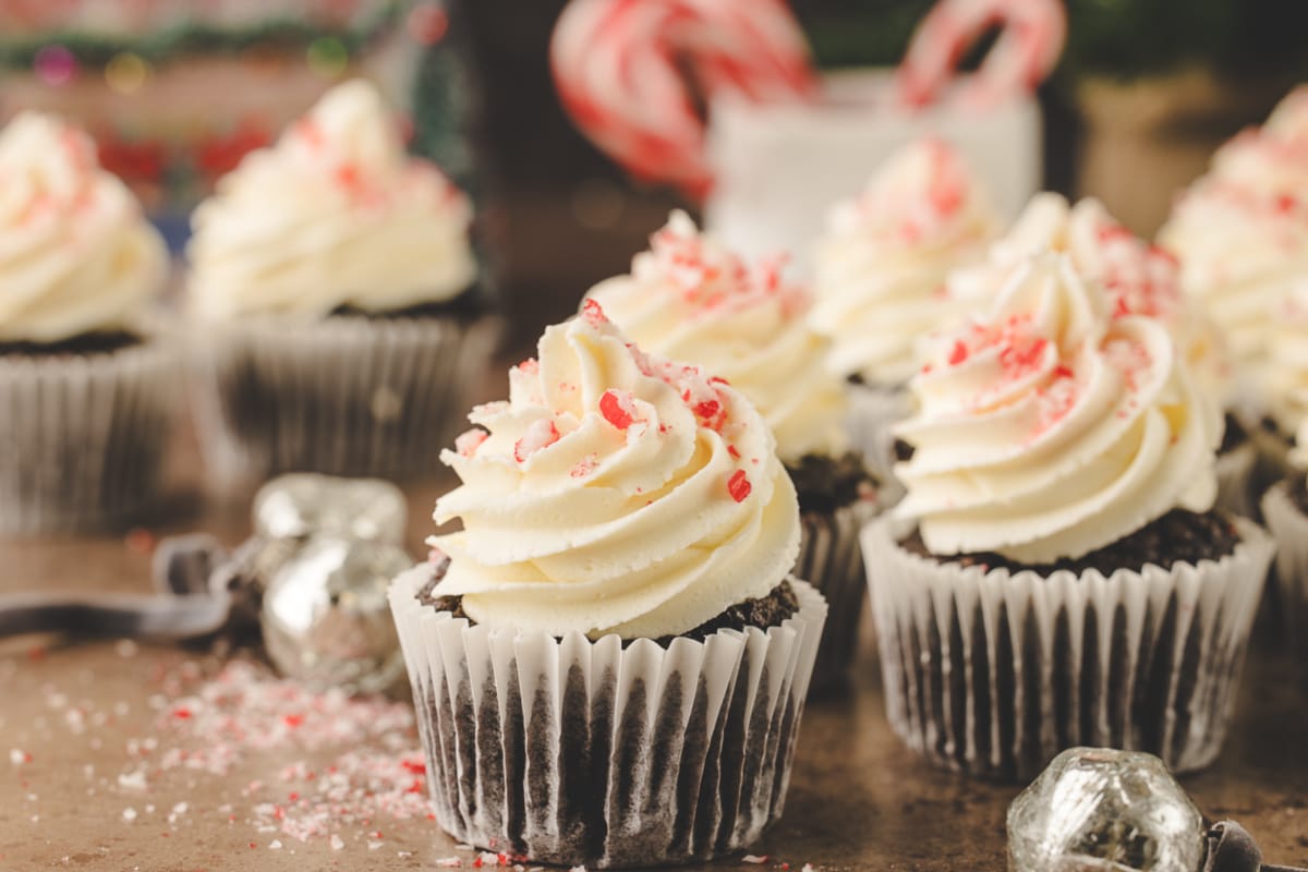 Chocolate cupcakes with white frosting and crushed peppermint, surrounded by holiday decorations and candy canes in the background.
