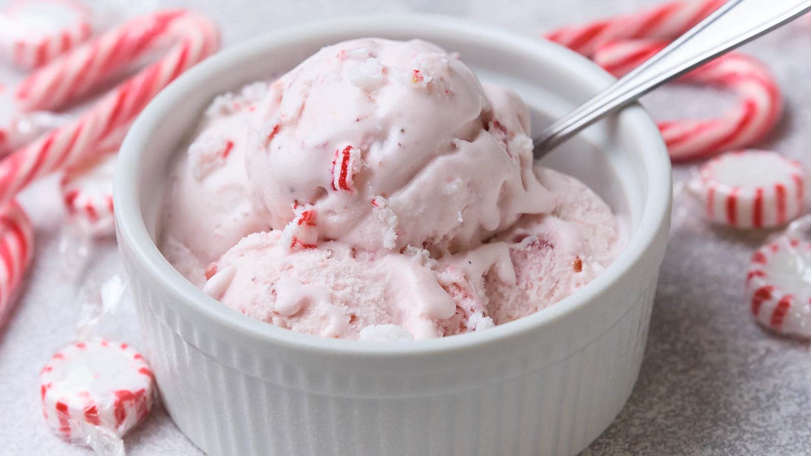 A white ramekin filled with scoops of peppermint ice cream, with a spoon inside. Peppermint candies and candy canes are visible in the background.