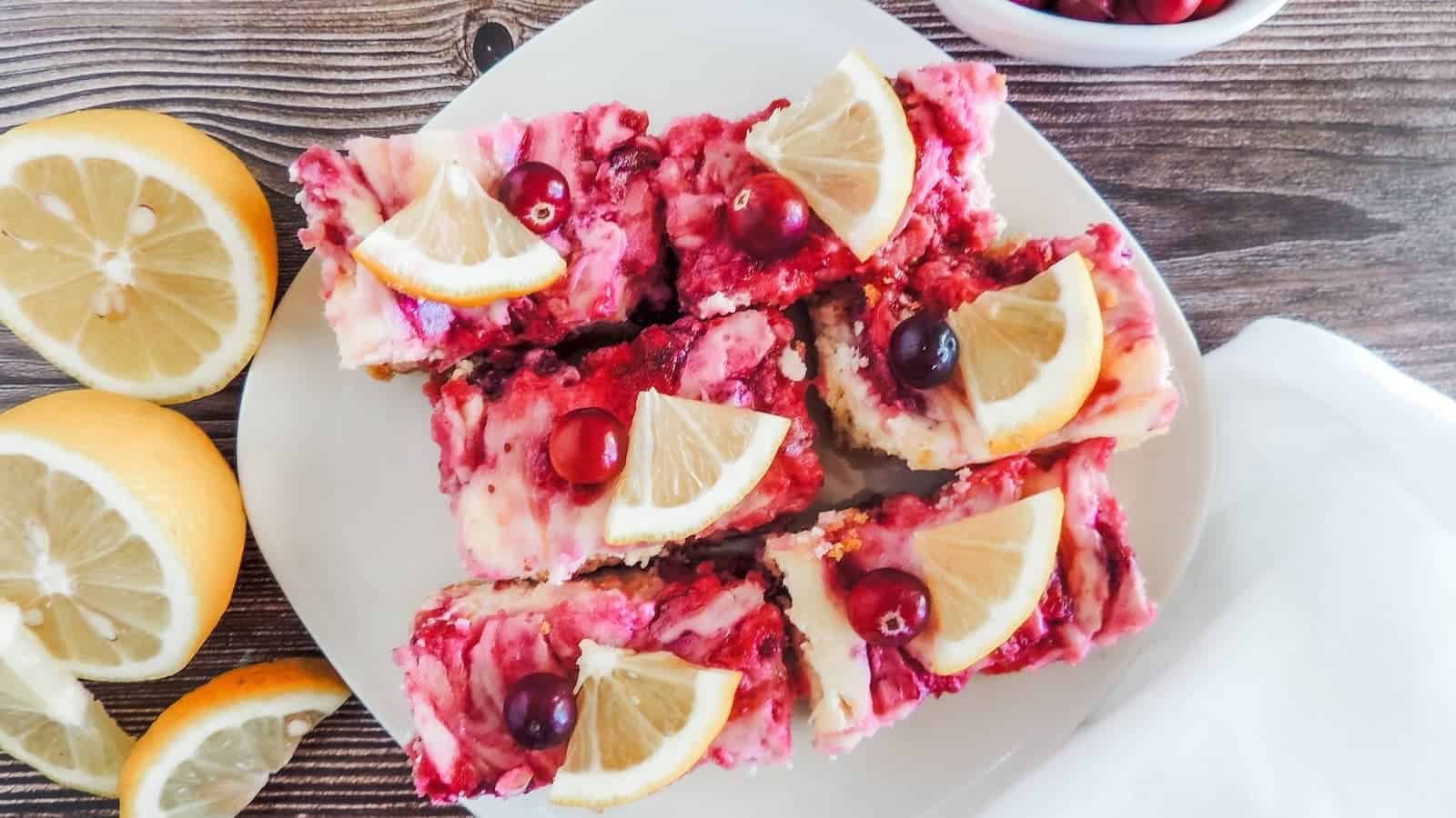 A plate of cranberry lemon cheesecake bars garnished with lemon slices and cranberries, set on a wooden surface with sliced lemons nearby.
