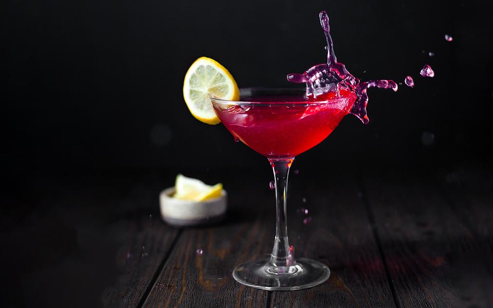A pink cocktail in a coupe glass garnished with a lemon slice, with a splash of liquid and lemon wedges in a bowl in the background on a dark wooden surface.