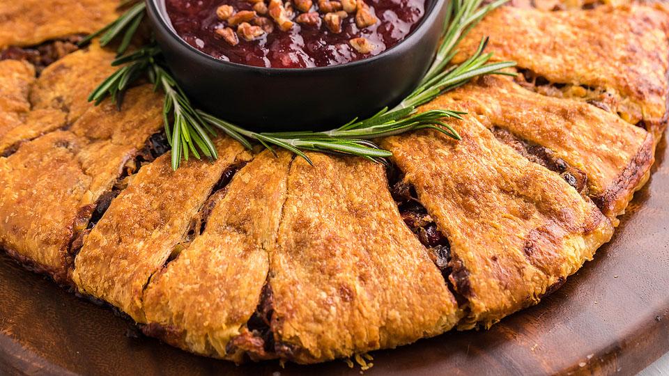A round baked pastry cut into slices surrounds a bowl of dark red dipping sauce, garnished with sprigs of rosemary on a wooden surface.