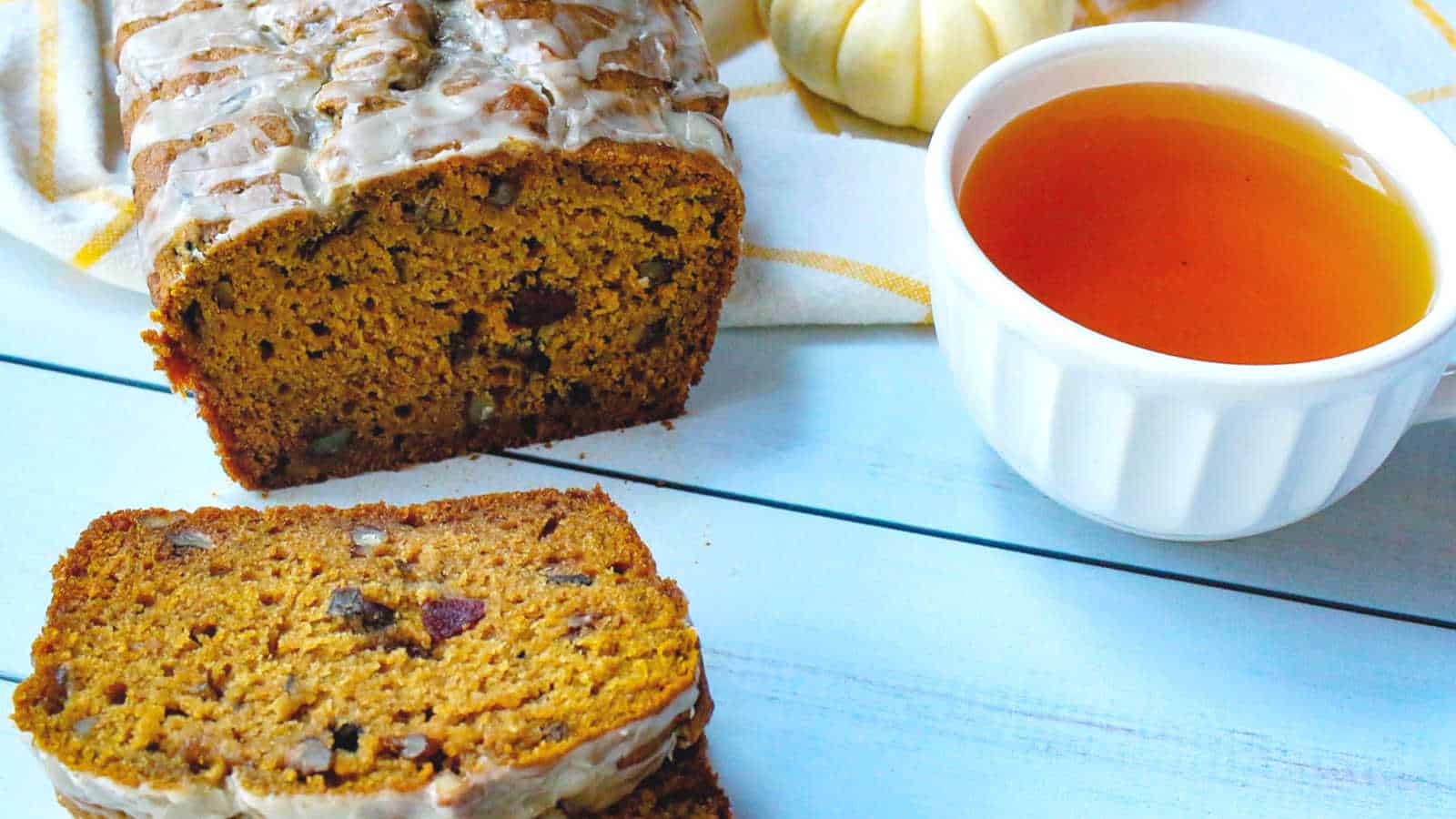 A loaf of pumpkin bread with icing sits next to two slices and a cup of tea on a light blue wooden surface. A small white pumpkin is in the background.