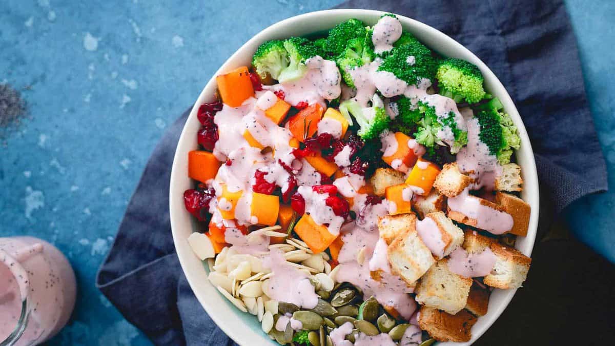 A salad bowl with broccoli, croutons, cubed squash, dried cranberries, sliced almonds, pumpkin seeds, and pink dressing on a blue surface.