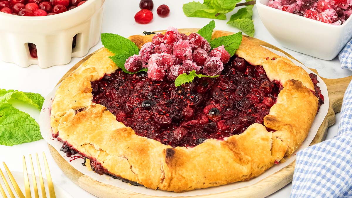 A rustic berry galette topped with sugared cranberries and mint leaves, surrounded by fresh berries and mint on a white table.
