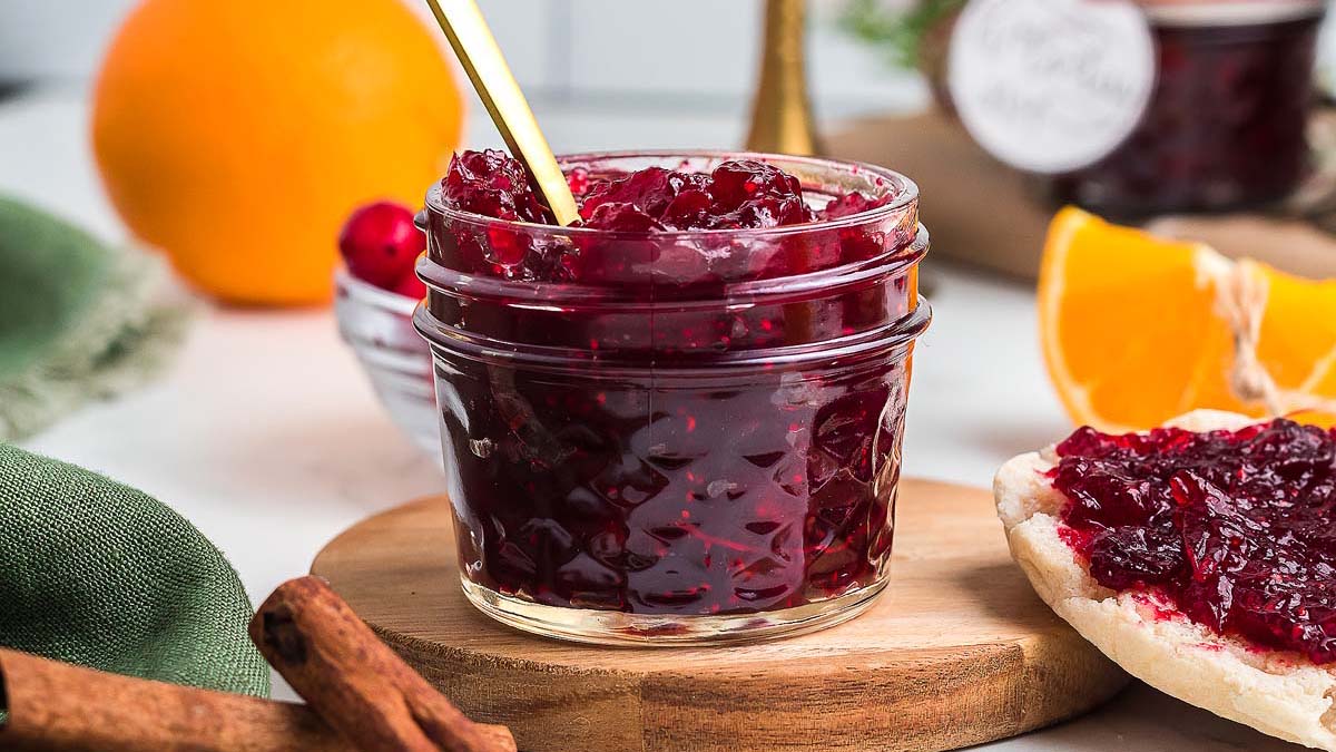 A glass jar filled with cranberry jam sits on a wooden board with a gold spoon inside. Nearby are orange slices, a cinnamon stick, and a piece of bread spread with jam.