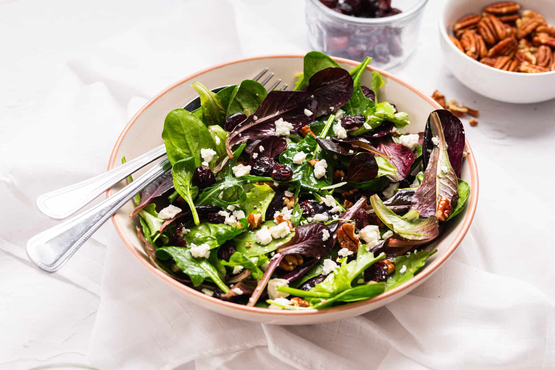 A bowl of mixed green salad with crumbled cheese, pecans, and dried cranberries, with a fork placed inside the bowl.