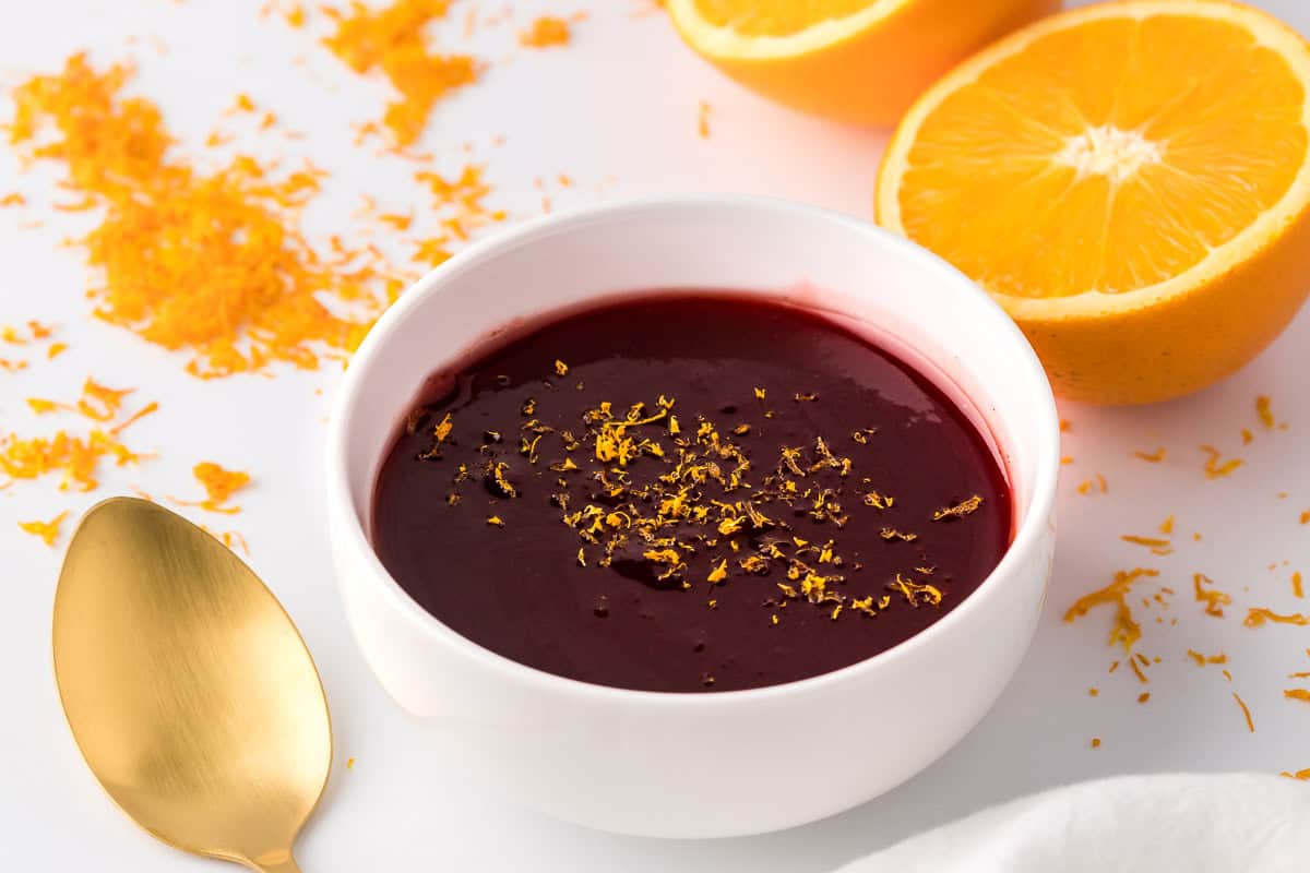 A white bowl filled with dark red sauce garnished with orange zest, set beside a gold spoon and a halved orange on a white surface.